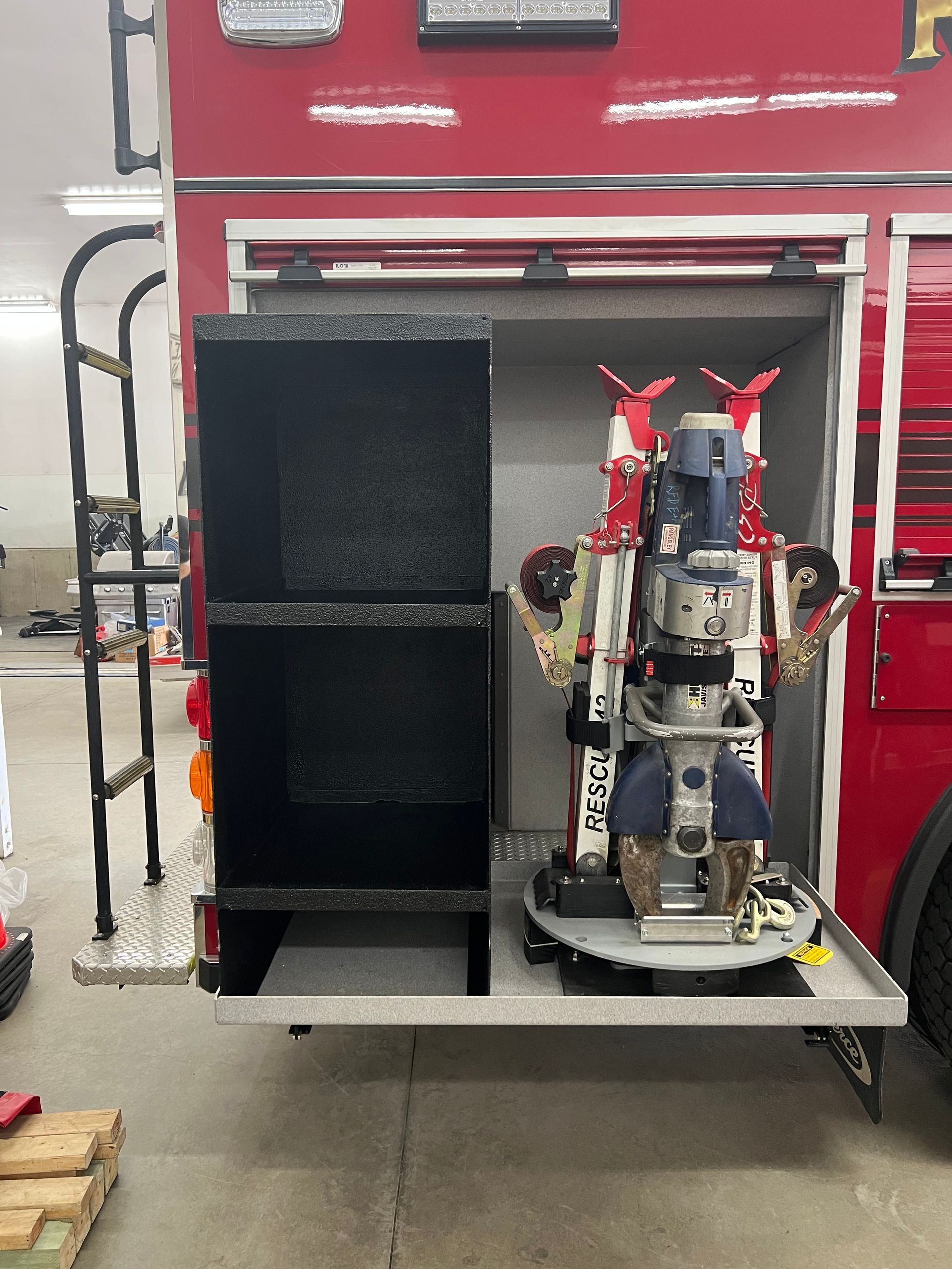 Open storage compartment on a red fire truck, containing two empty black shelves and specialized rescue tools on a mount.