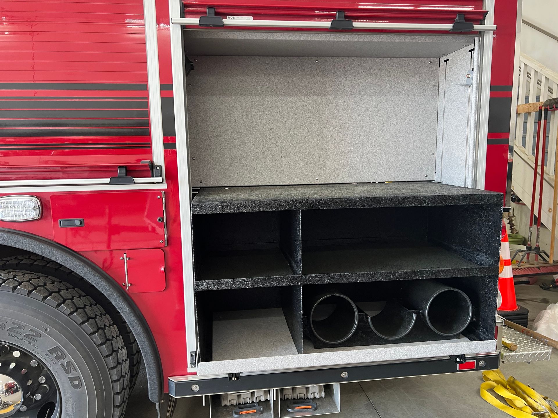 A side view of a red fire truck with an open storage compartment revealing interior shelving and equipment tubes.