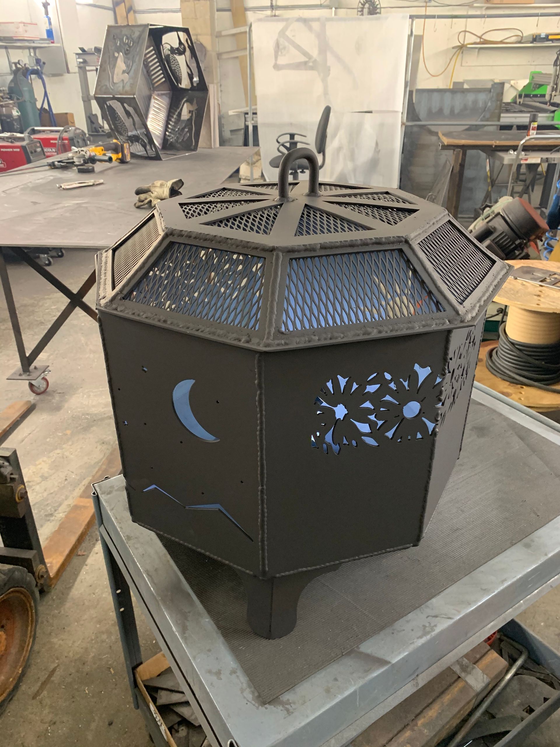 A dark, octagonal metal fire pit with cutout designs of a moon and flowers, sitting on a work table in a metal shop.