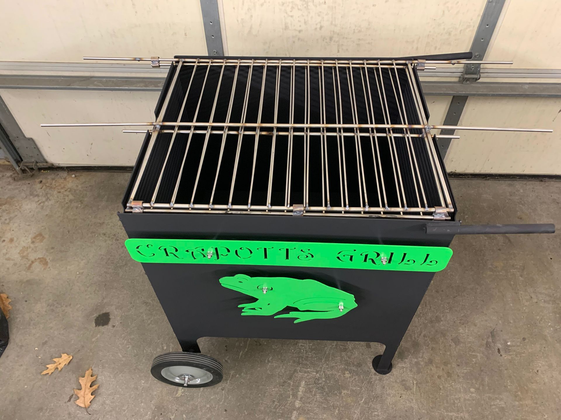 A black metal charcoal grill with a green frog cutout and the name