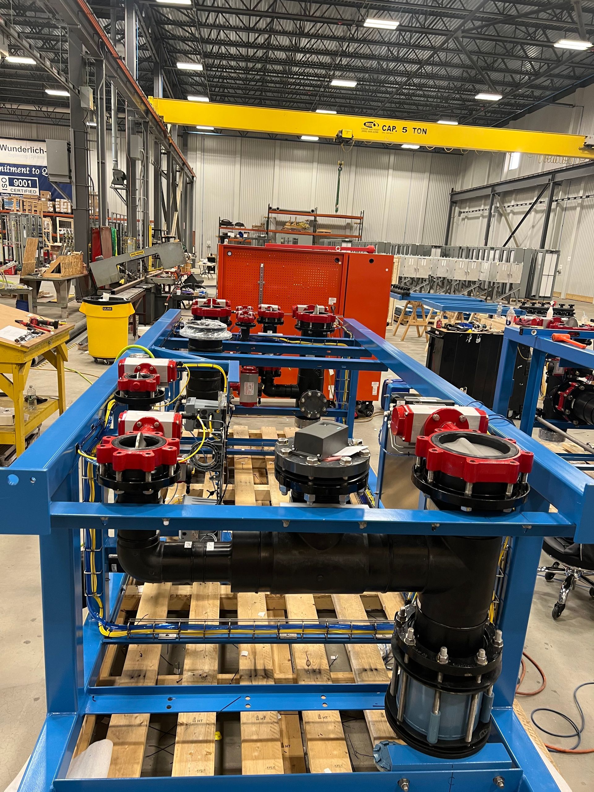 Industrial facility showing a blue metal frame containing black pipes and red valve components on a wooden pallet.