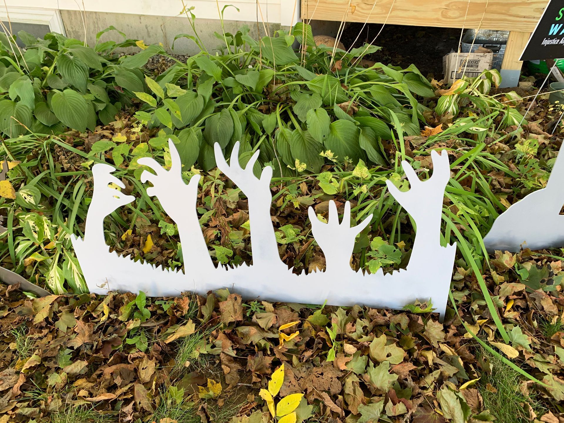 White cutout of zombie hands reaching up from a garden bed covered in autumn leaves.
