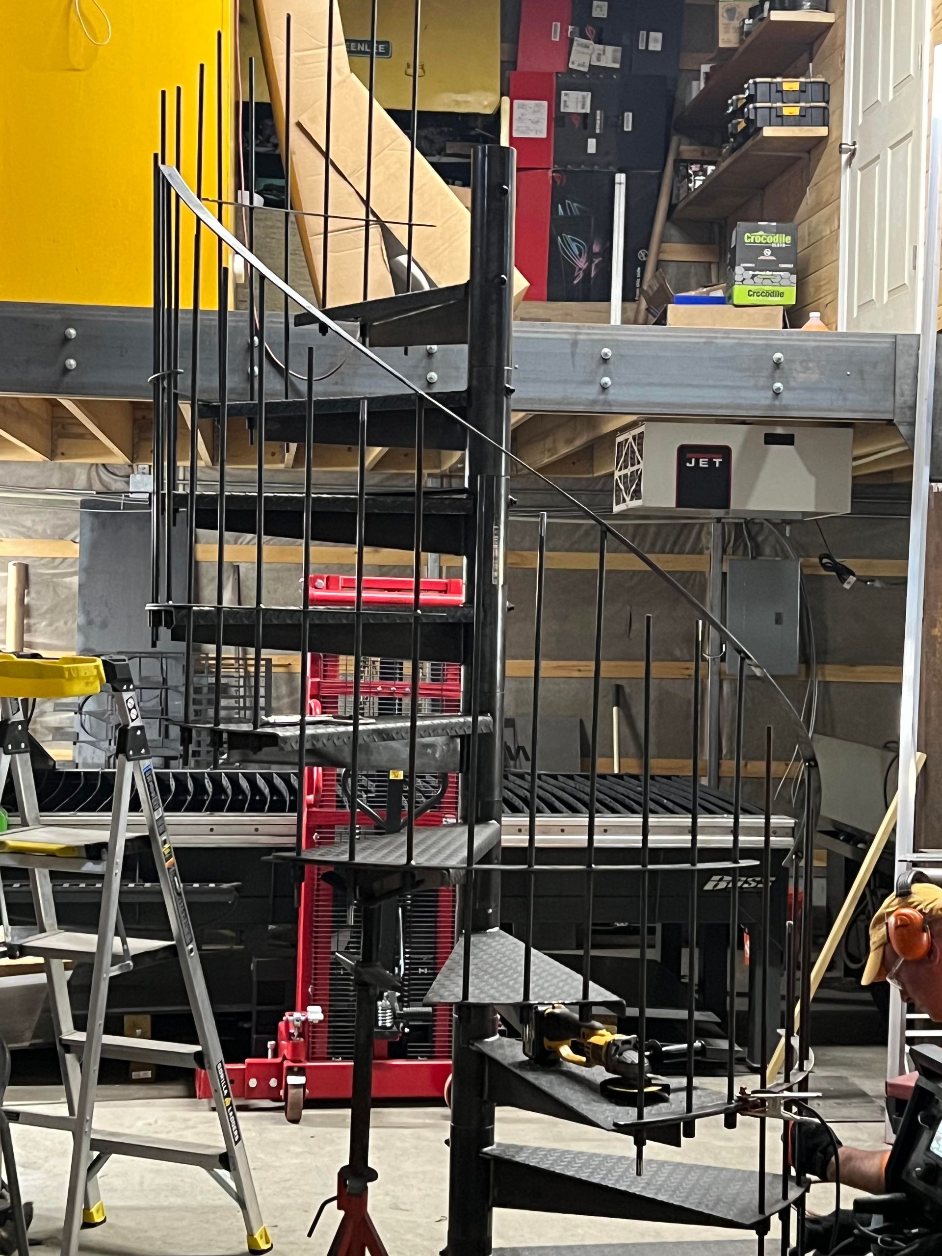A black metal spiral staircase being assembled in a workshop, with tools and construction equipment nearby.