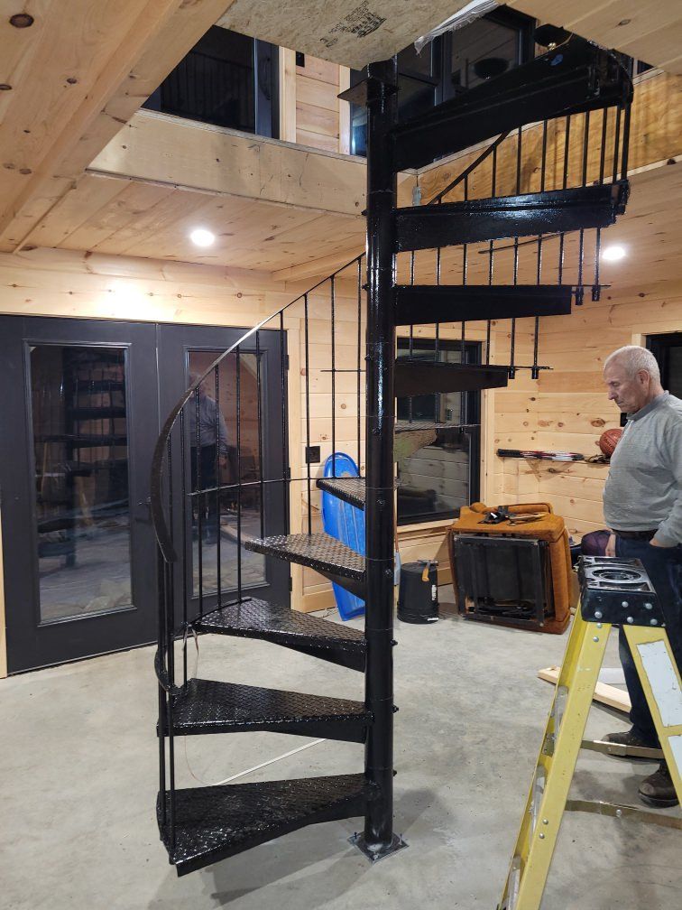 A black metal spiral staircase being installed in a room with light wood walls, as a person stands nearby on a ladder.