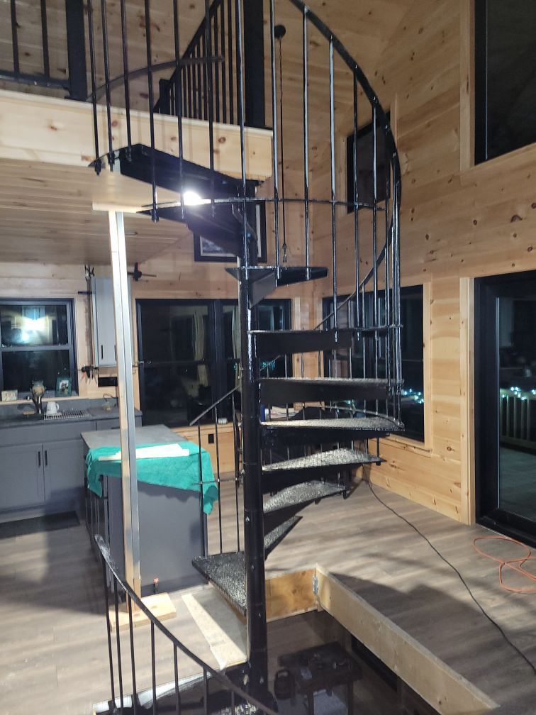 A black metal spiral staircase stands in a cabin-style room with light wood walls and flooring.
