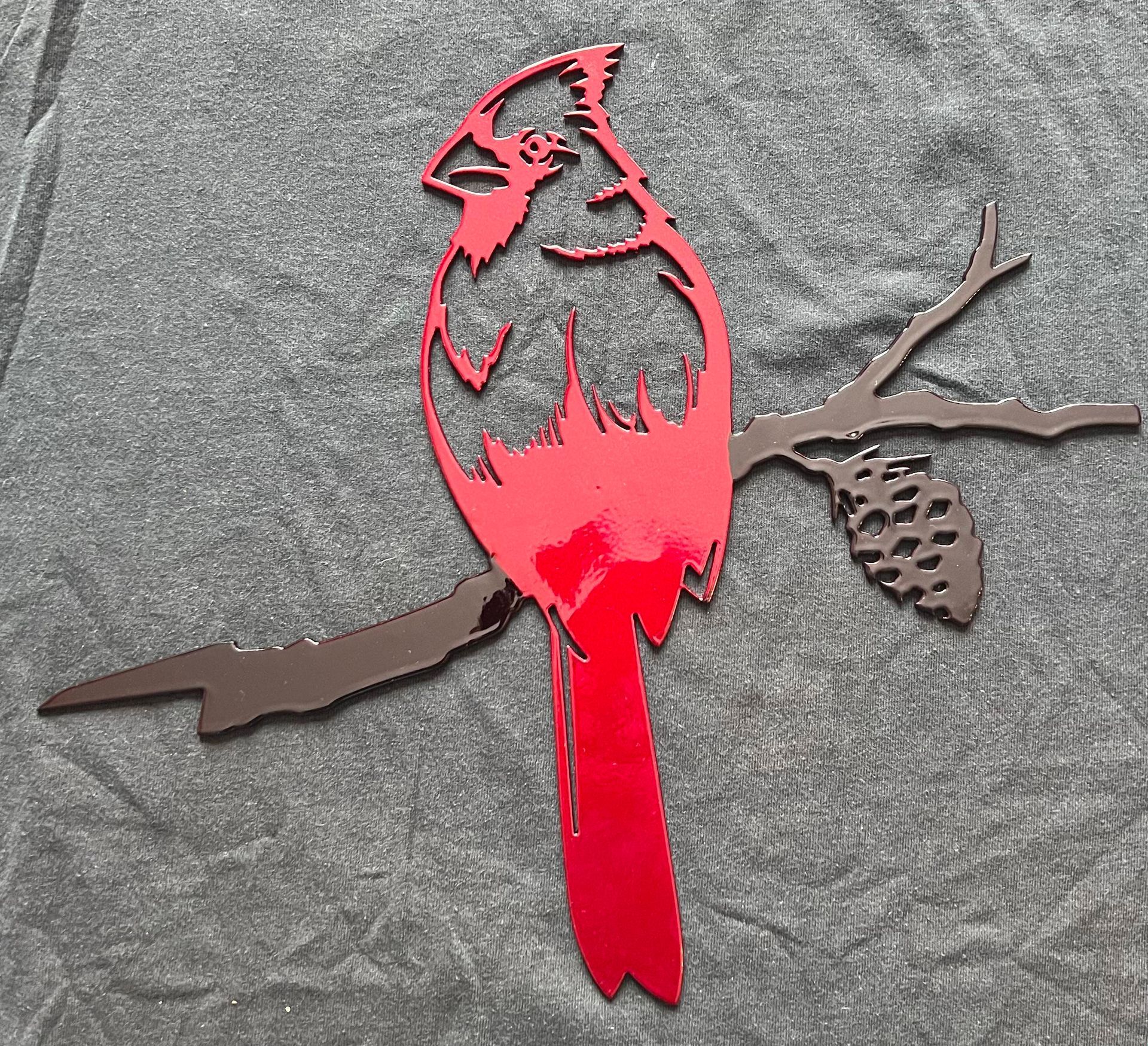 A bright red metal cardinal perched on a brown branch with a pinecone, set against a grey fabric background.
