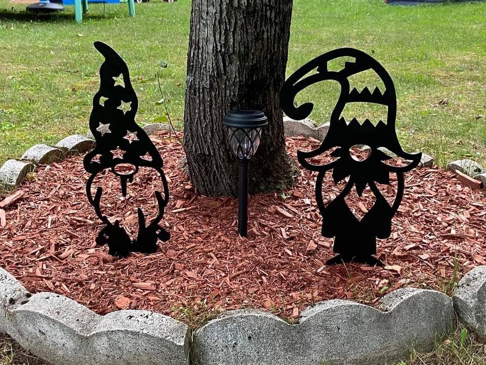 Two black metal gnome silhouettes stand in a mulched garden bed around a tree, with a small solar light between them.