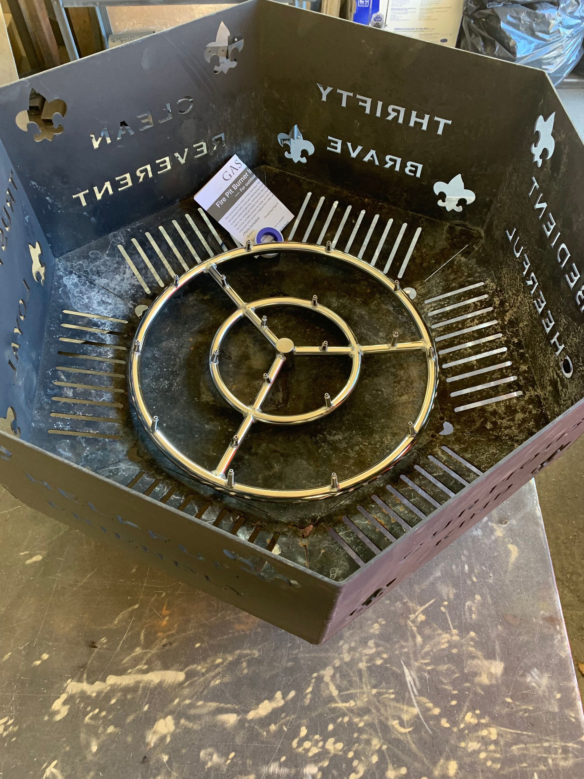 A hexagonal metal fire pit featuring a circular gas burner ring and cutouts of the Scout Law values.