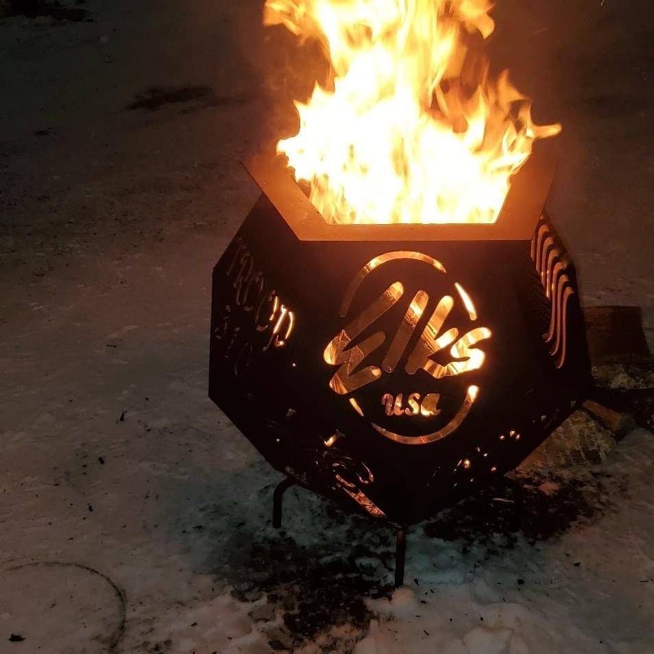 A black, metal fire pit shaped like a pentagon glows with bright flames, featuring the