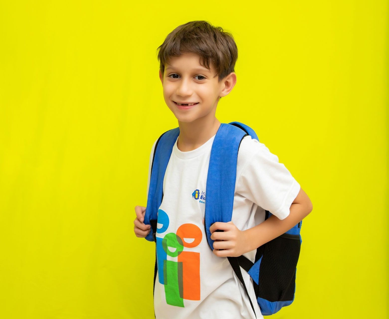 Um garoto vestindo uma camisa branca e uma mochila azul está parado em frente a uma parede amarela.