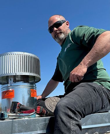 Man in green shirt, sunglasses on rooftop, installing a silver chimney cap.