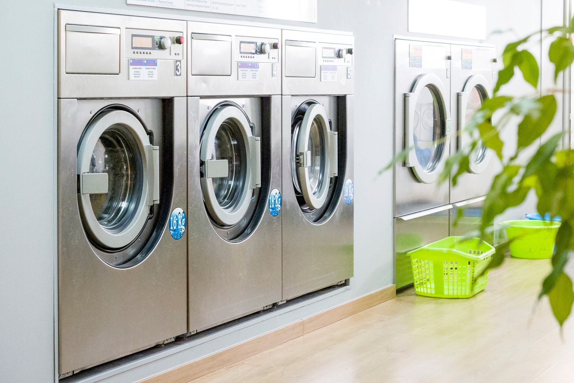 Row of washing machines in a laundromat, stainless steel, with green laundry baskets and plants.