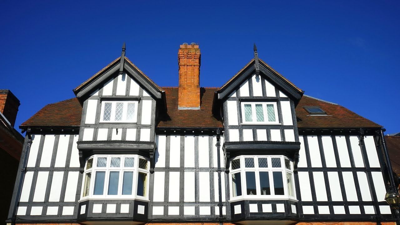 Tudor-style house with white and black beams, bay windows, and a brown roof against a clear blue sky.