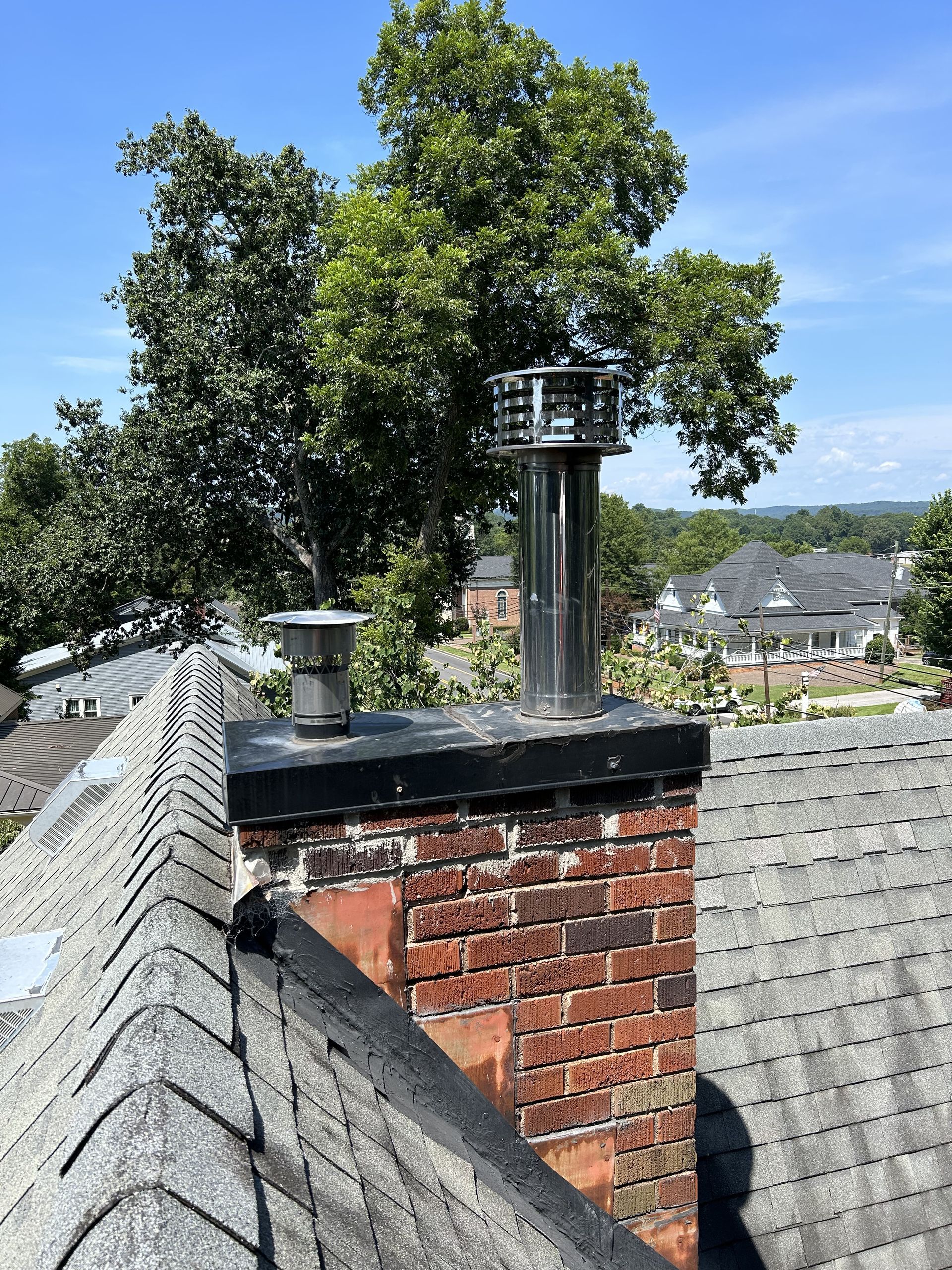 Brick chimney with metal vent on a shingled rooftop, with trees and houses in the background.