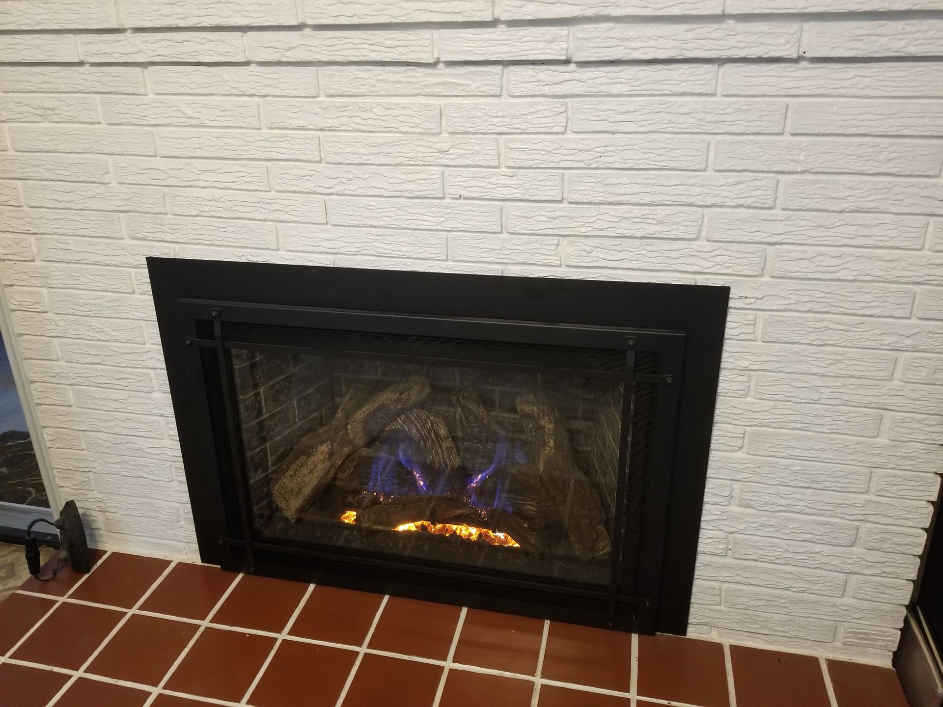 Lit fireplace with orange flames in a black surround set into a white brick wall above terracotta tile flooring