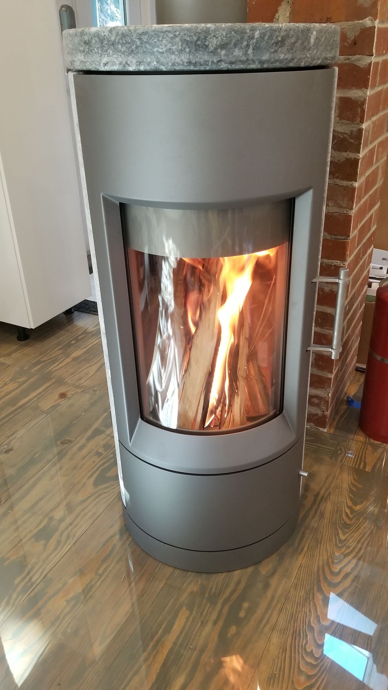 Gray cylindrical wood-burning stove with visible flames and logs inside a brick-lined room