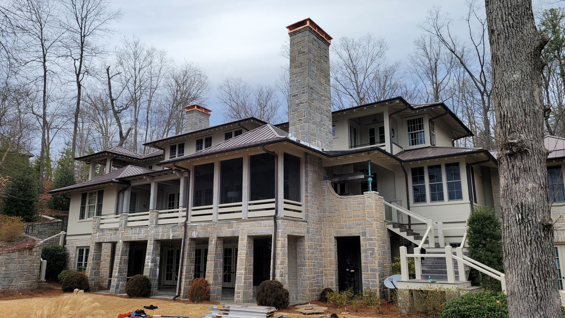 Large stone and wood house with a tall chimney and screened porches in a wooded area