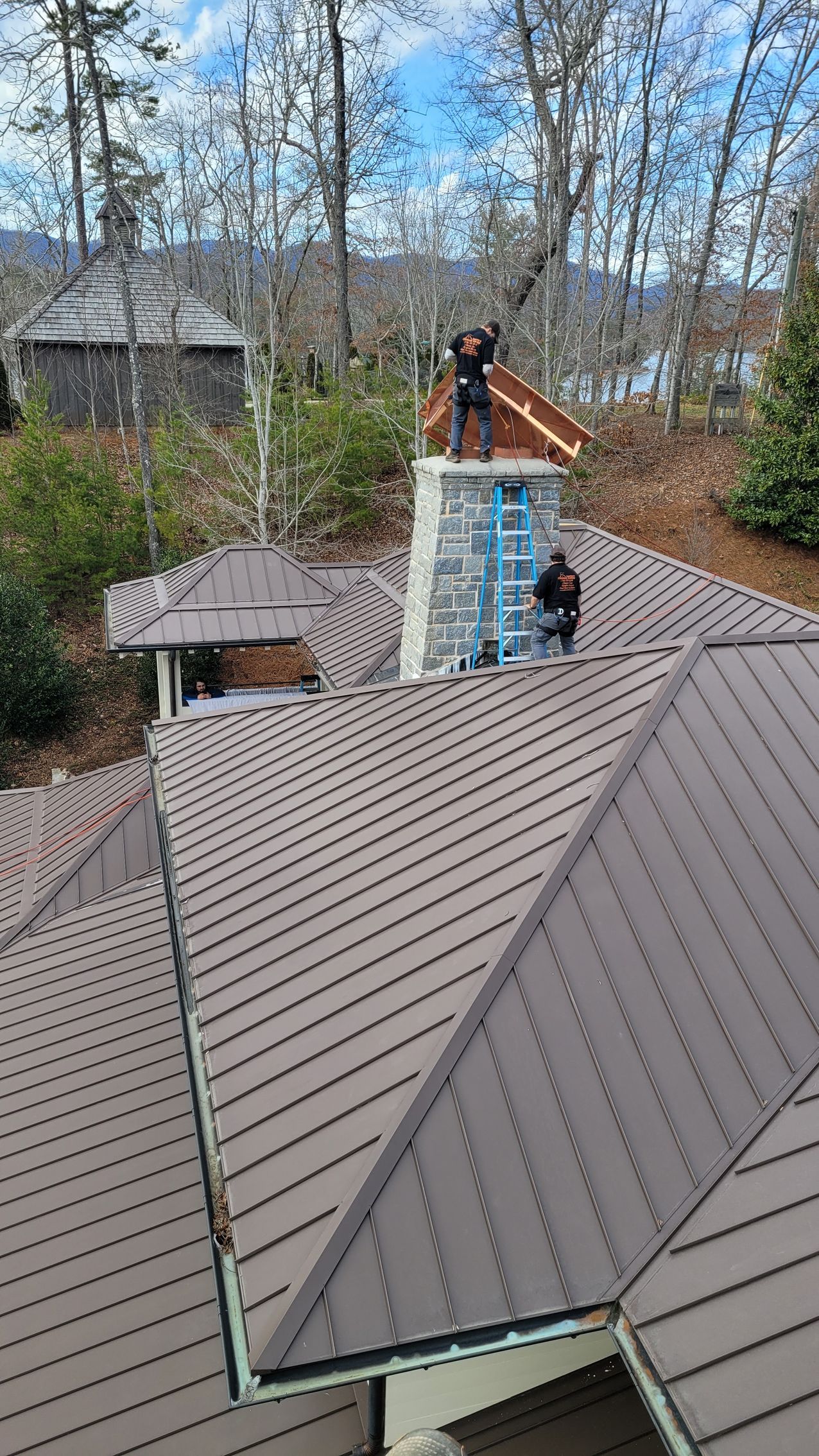 Roof workers on a gray shingled house beside a chimney and leafless trees