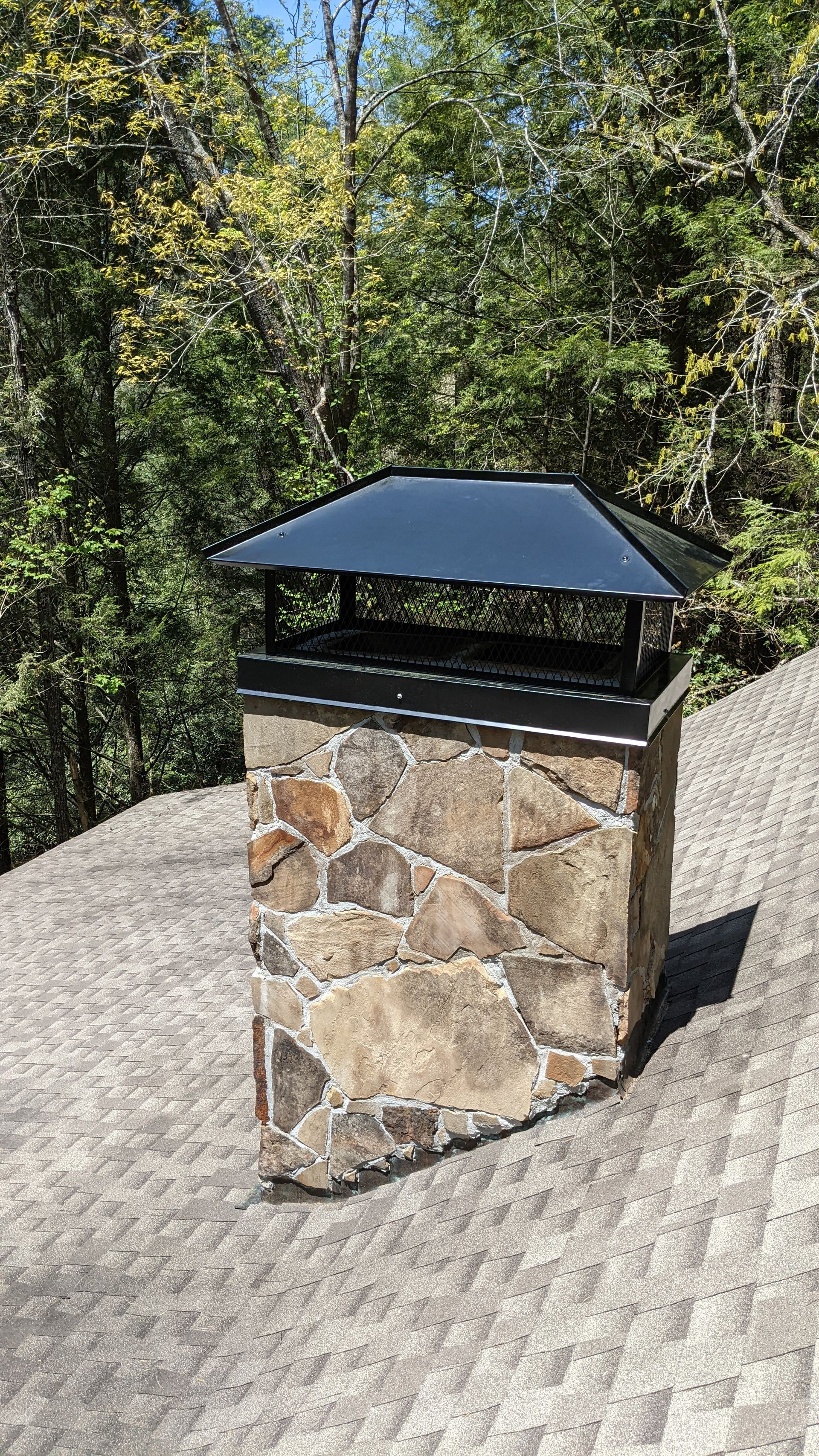 Stone chimney with black metal cap on a shingled roof, with trees in the background
