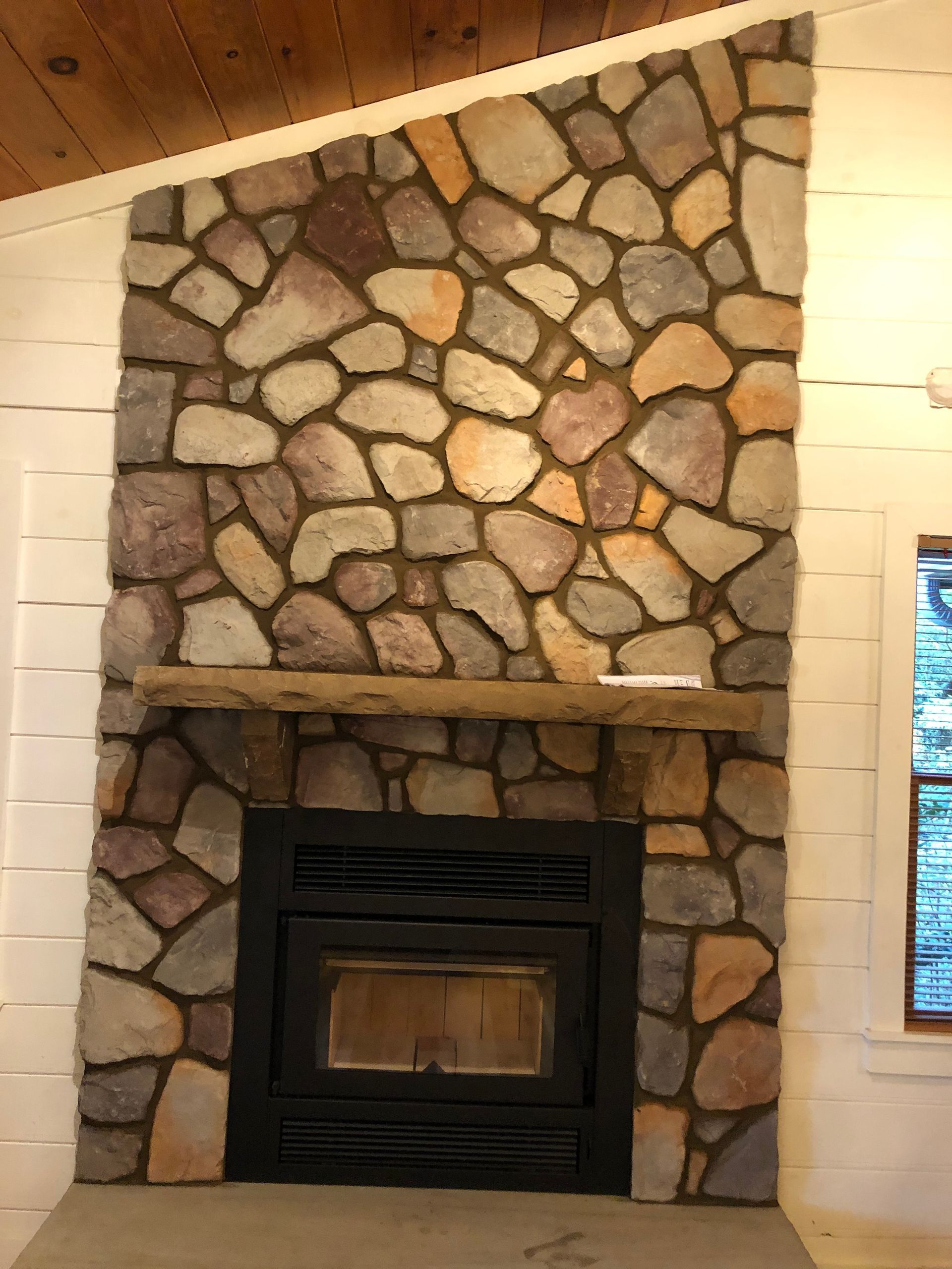 Stone fireplace with black wood stove insert and slanted mantel in a wood-paneled room
