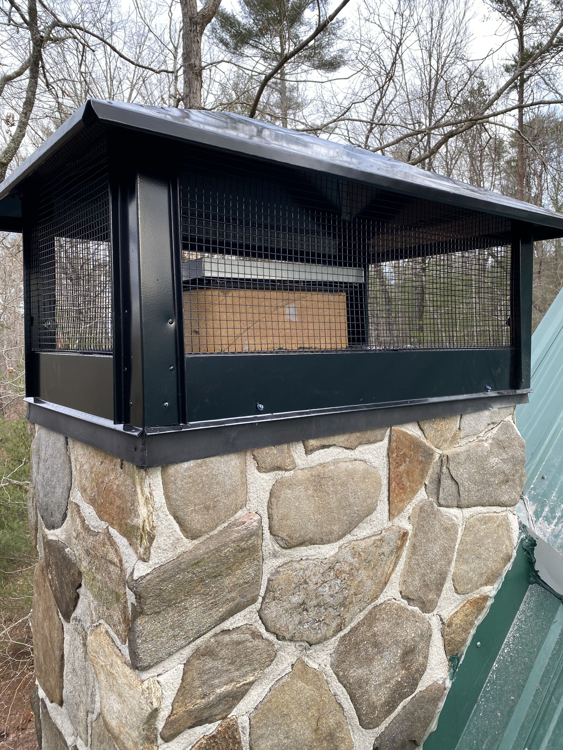 Stone outdoor fireplace with black metal hood and chimney, covered by a wire mesh screen