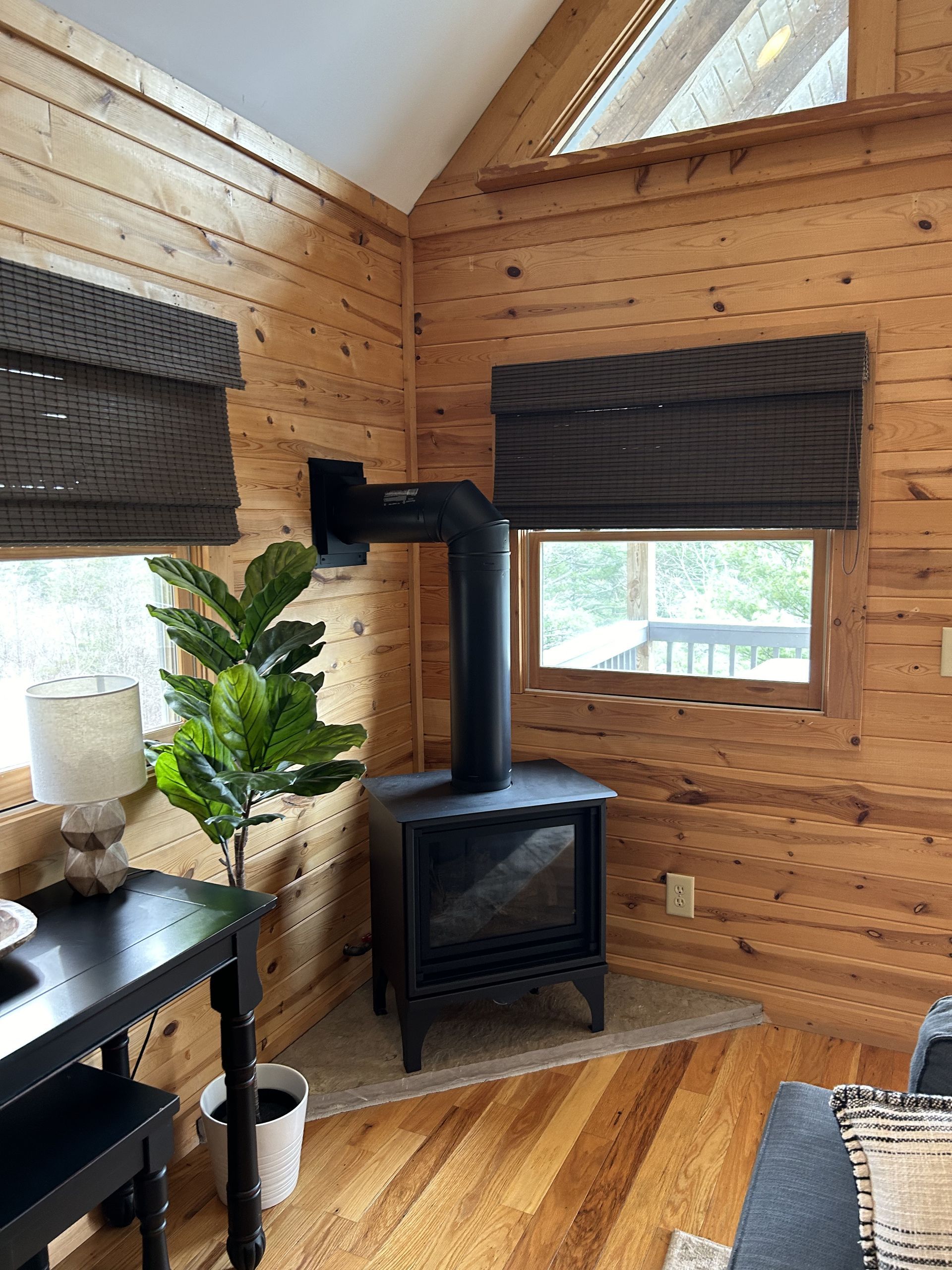 Wood-paneled cabin corner with black wood stove, potted plant, and windows with dark blinds