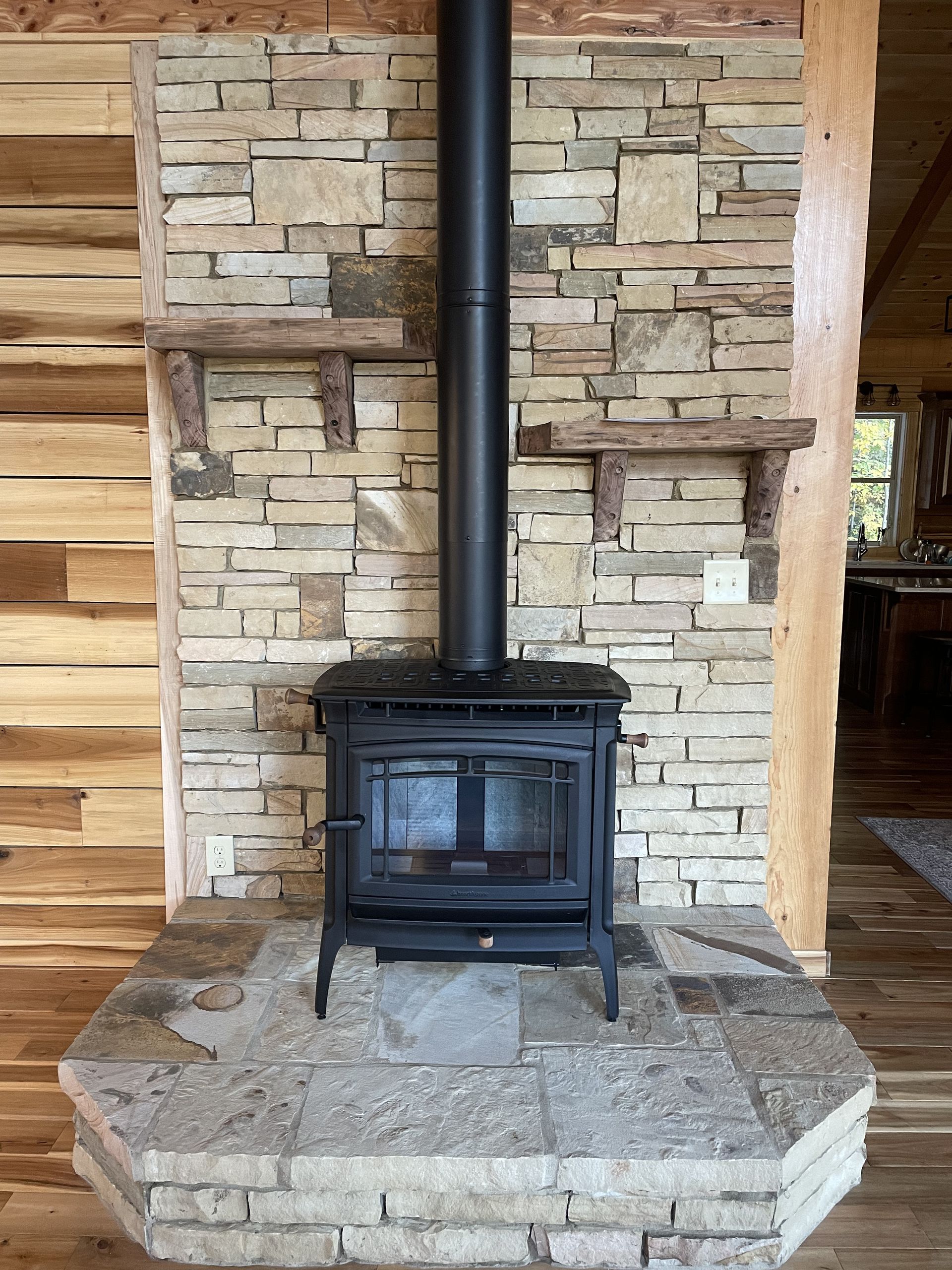 Black wood stove centered on a stone hearth in front of a rustic stone fireplace and wooden beams