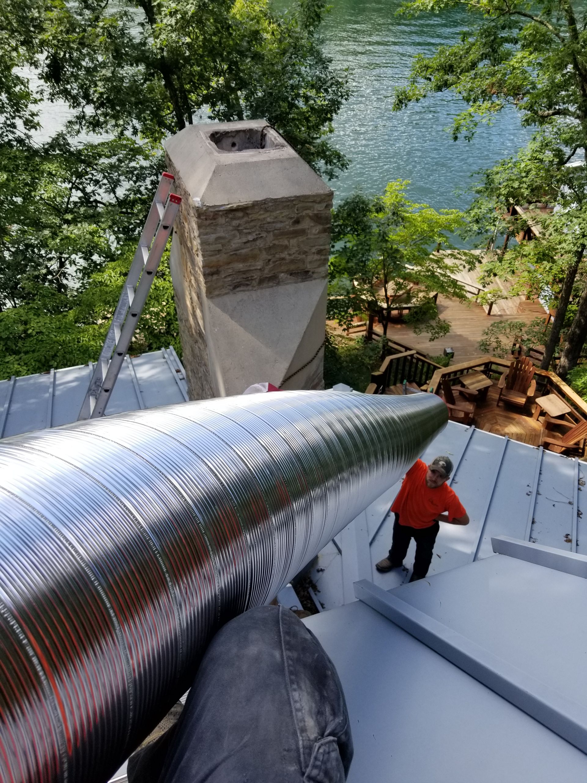 Worker on a roof installing silver insulation near a river, with a ladder and chimney nearby.