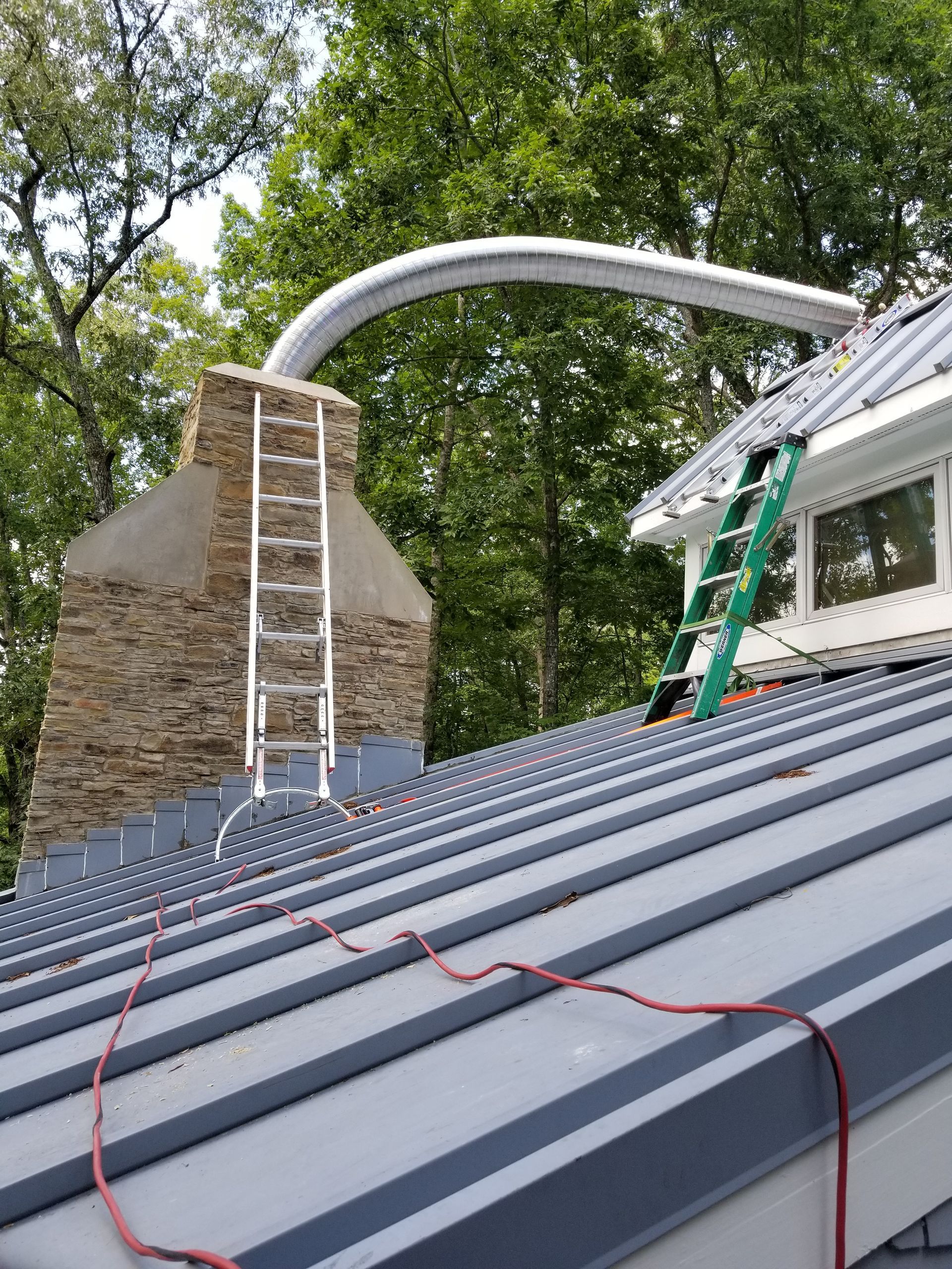 Workers on a steep metal roof beside a stone chimney, with ladders and green roof panels.