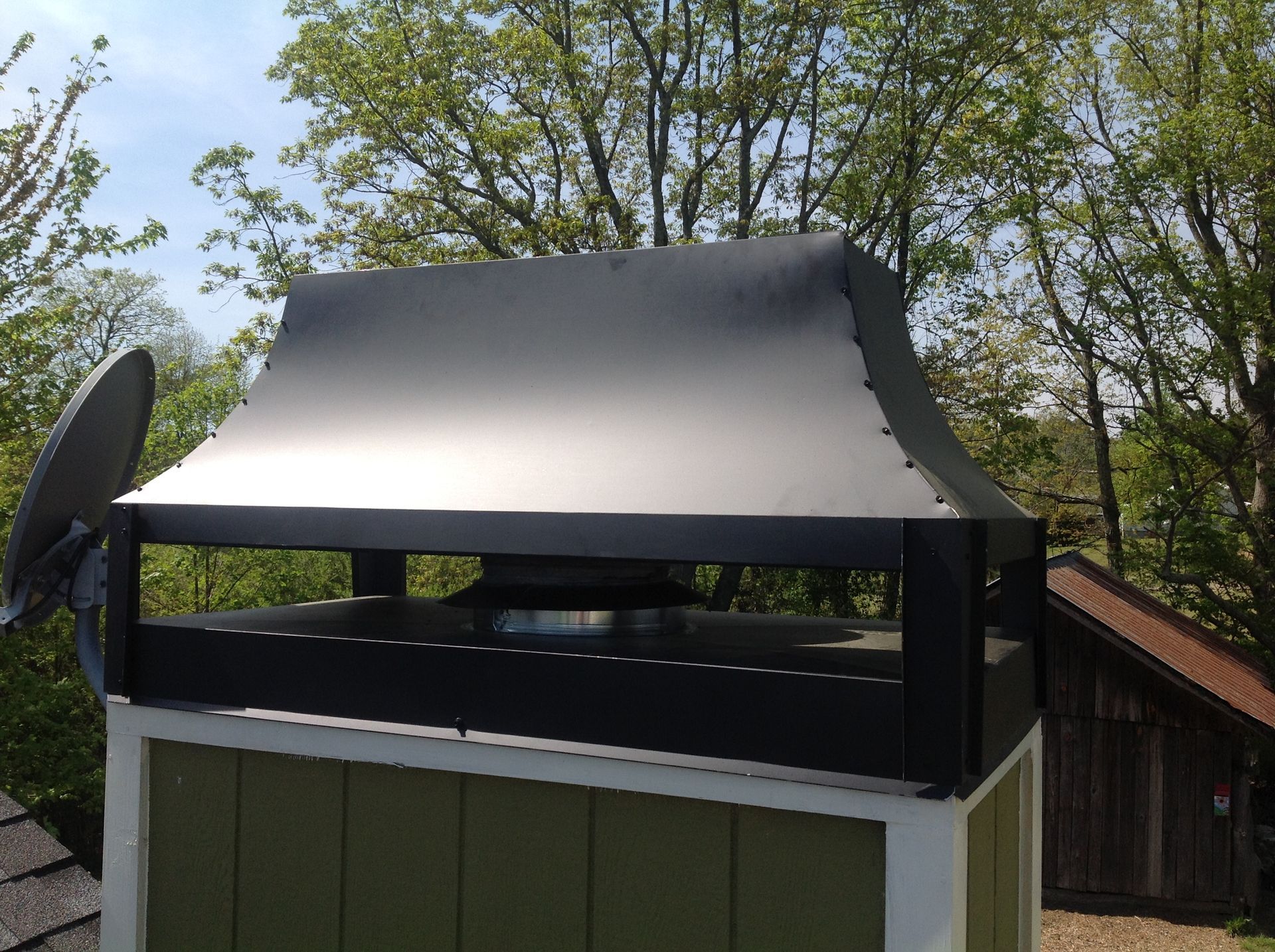 Black roof vent or chimney cap on a green shed, with trees in the background