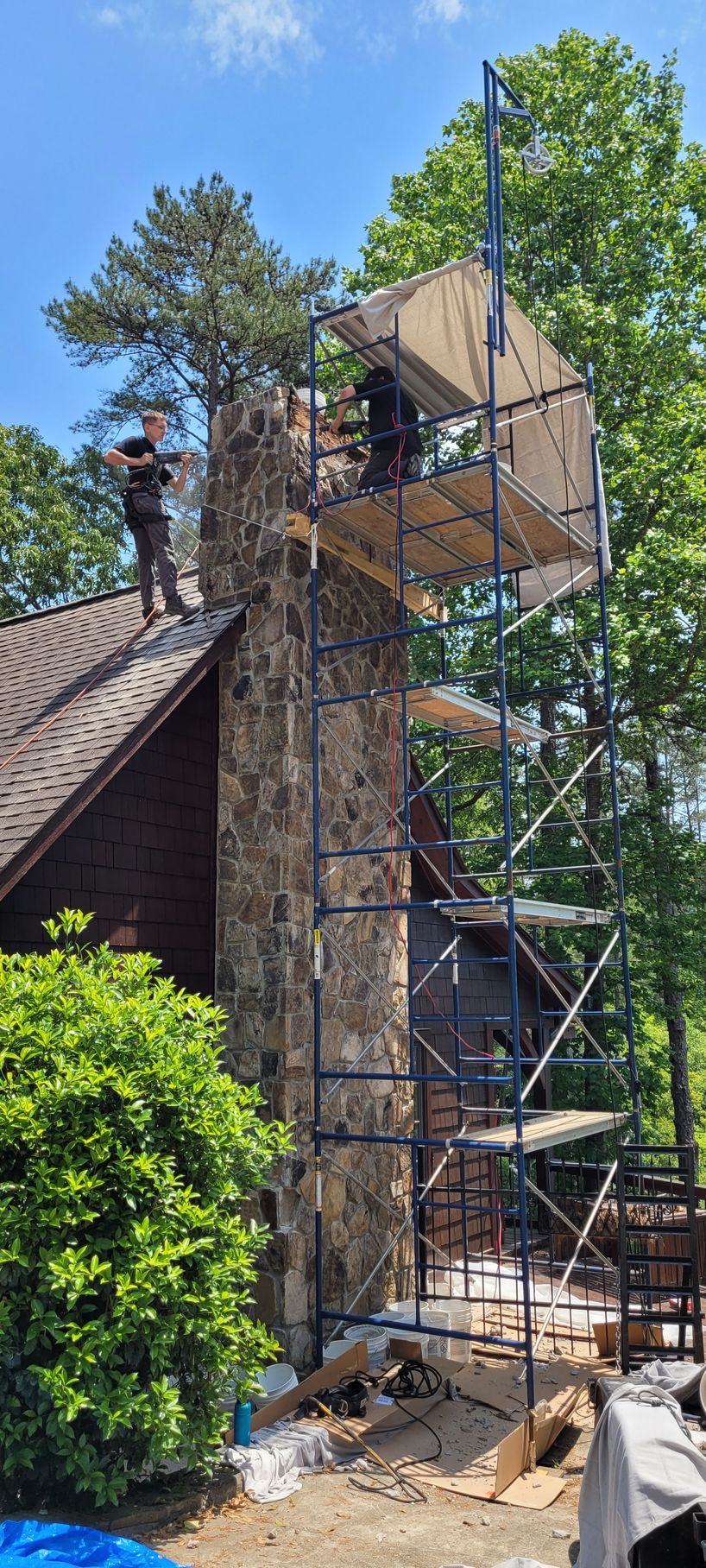 Scaffolding beside a stone chimney under repair, with a worker on top against a blue sky.