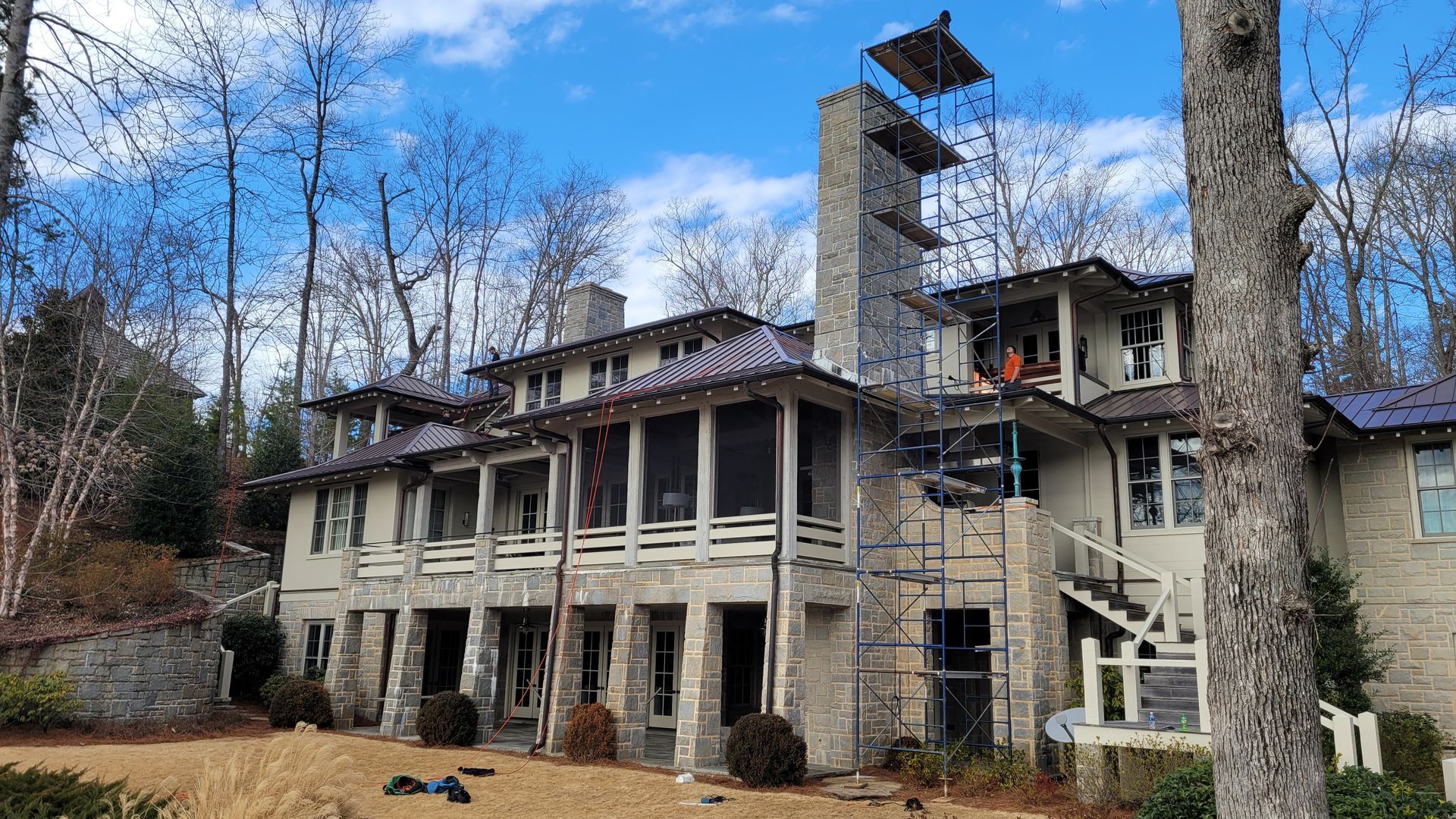 Large stone house with tall chimney and balconies in a wooded area under a partly cloudy sky
