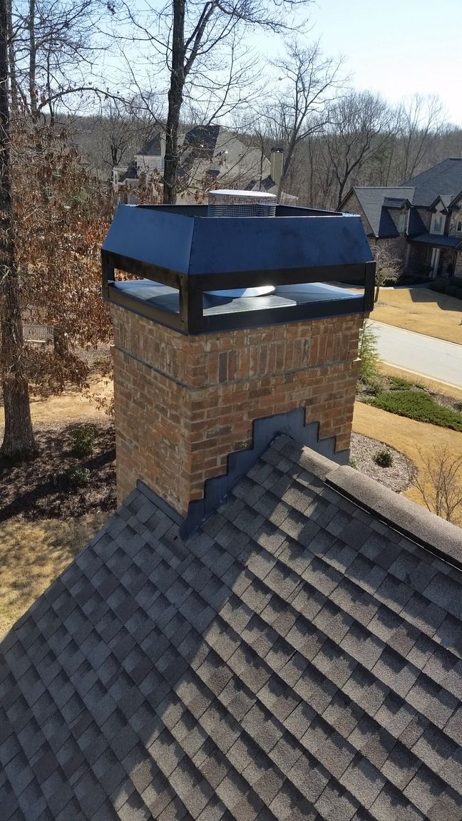 Brick chimney with black metal cap on a shingled roof, trees and houses in the background
