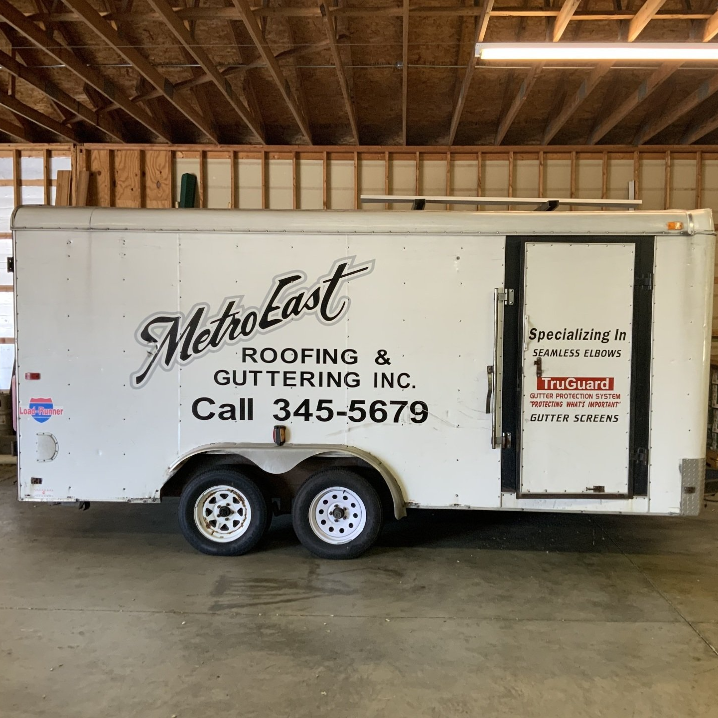 White roofing company trailer in a garage, labeled “MetroLast Roofing & Guttering Inc.” with phone number 345-5679