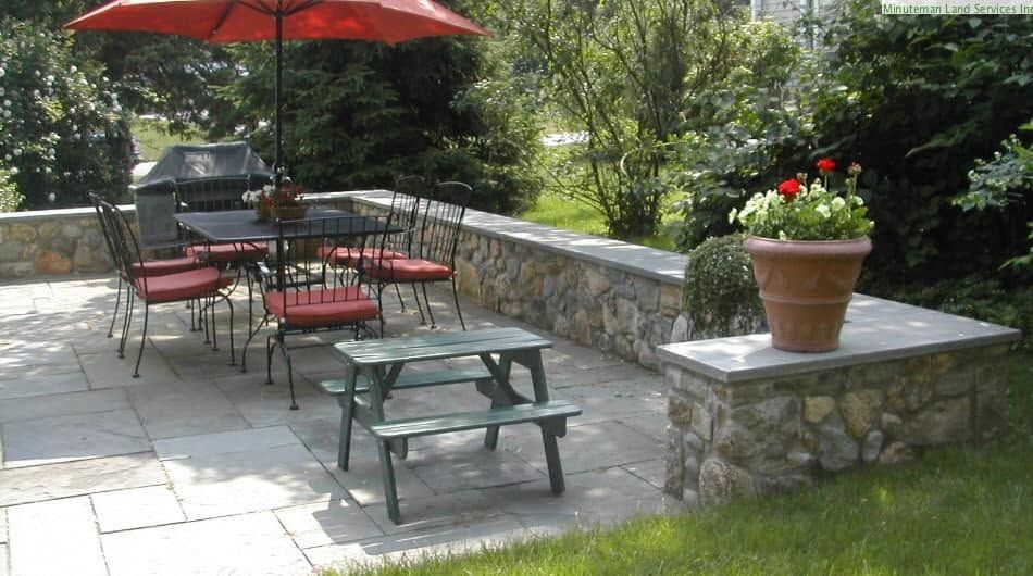 Patio with red umbrella, picnic table, chairs, stone wall, and potted flowers in a garden setting
