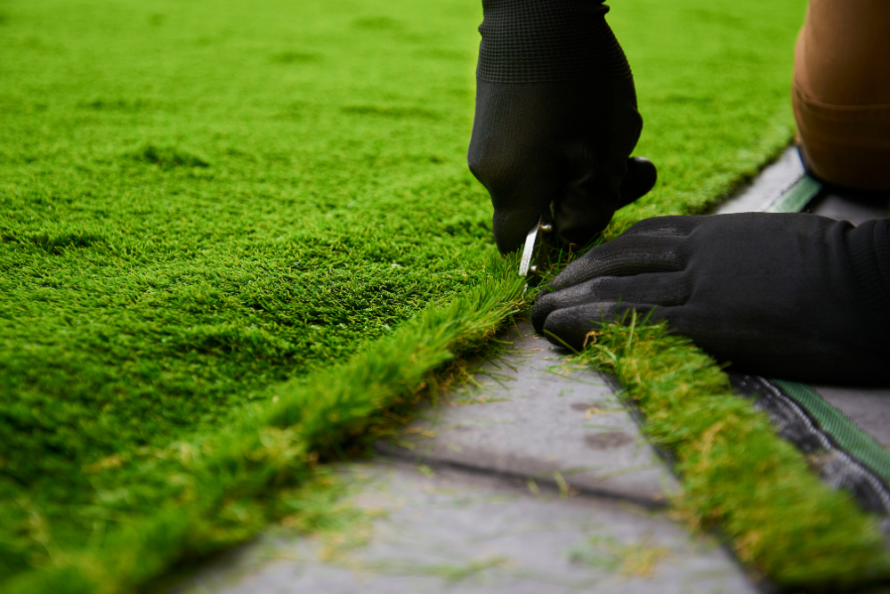 Gloved hands trimming grass along a stone edge with a small blade tool