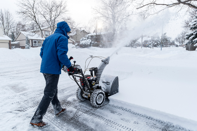 Person in blue jacket clearing snow with a snowblower on a snowy residential street.