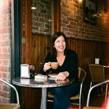 A smiling person sits at a small, round metal table in a brick-walled cafe, holding a coffee cup.