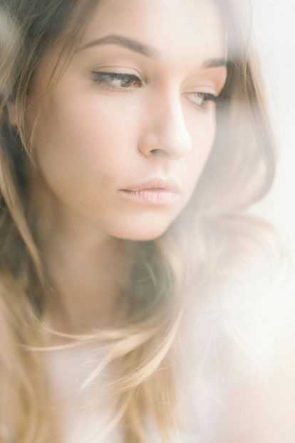 Hands hold a small mirror reflecting a partial view of a face, focused on an eye and lips, in a soft-focus, sepia tone.