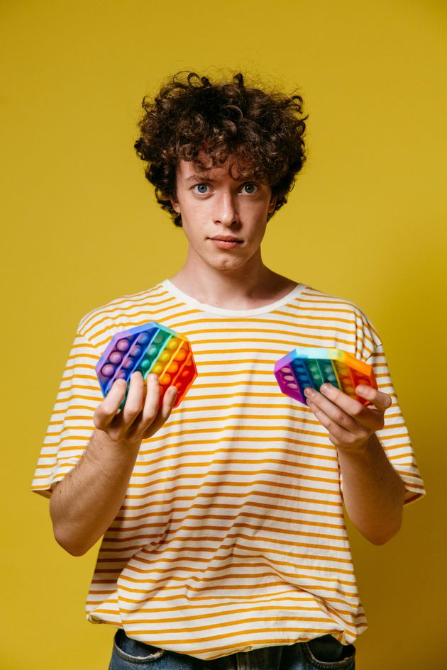 A person with curly hair wearing a yellow striped shirt holds two colorful, hexagonal, rainbow-patterned fidget poppers.