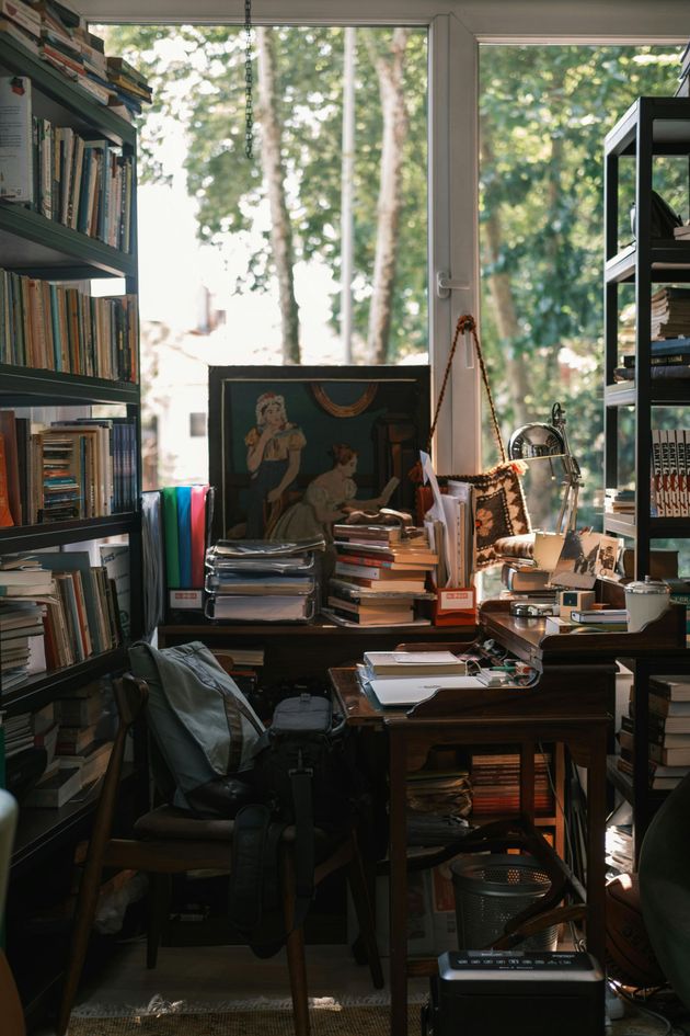 A cluttered home office with wooden desks, stacked books, shelving units, a large window, and a framed painting.