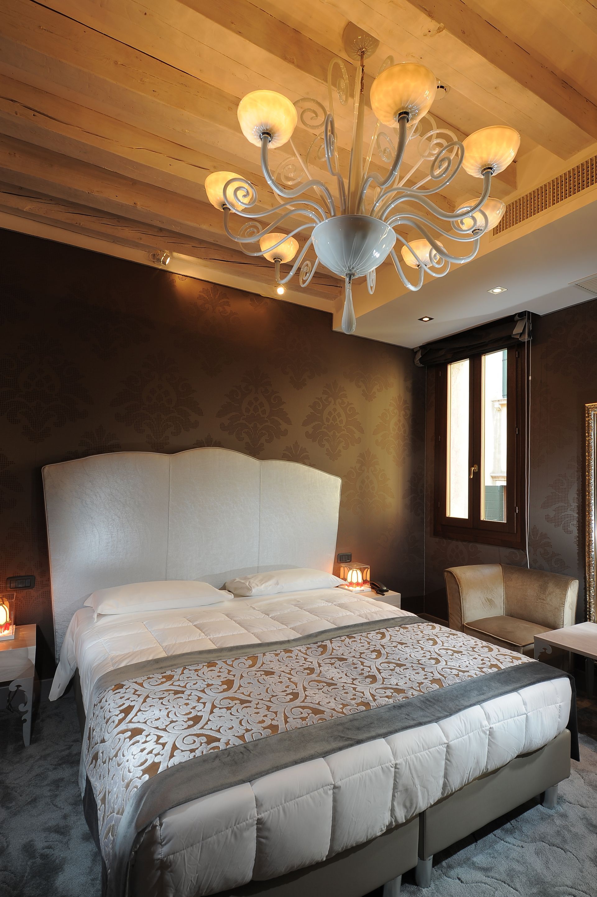 Cozy bedroom with white bed, patterned bedding, wood ceiling, chandelier, and a window seat