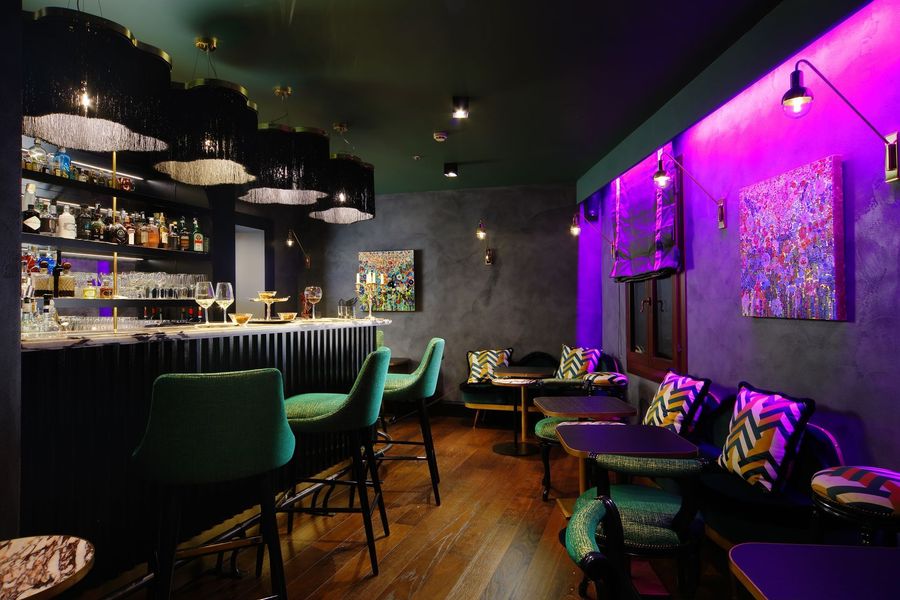 Dim neon-lit bar lounge with green chairs, shelves of bottles, and purple wall lighting