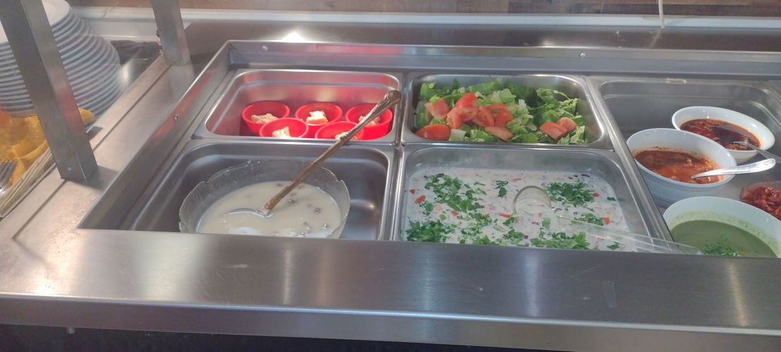 Food serving station with various dishes in metal containers. Red bell peppers, salads, and sauces visible.