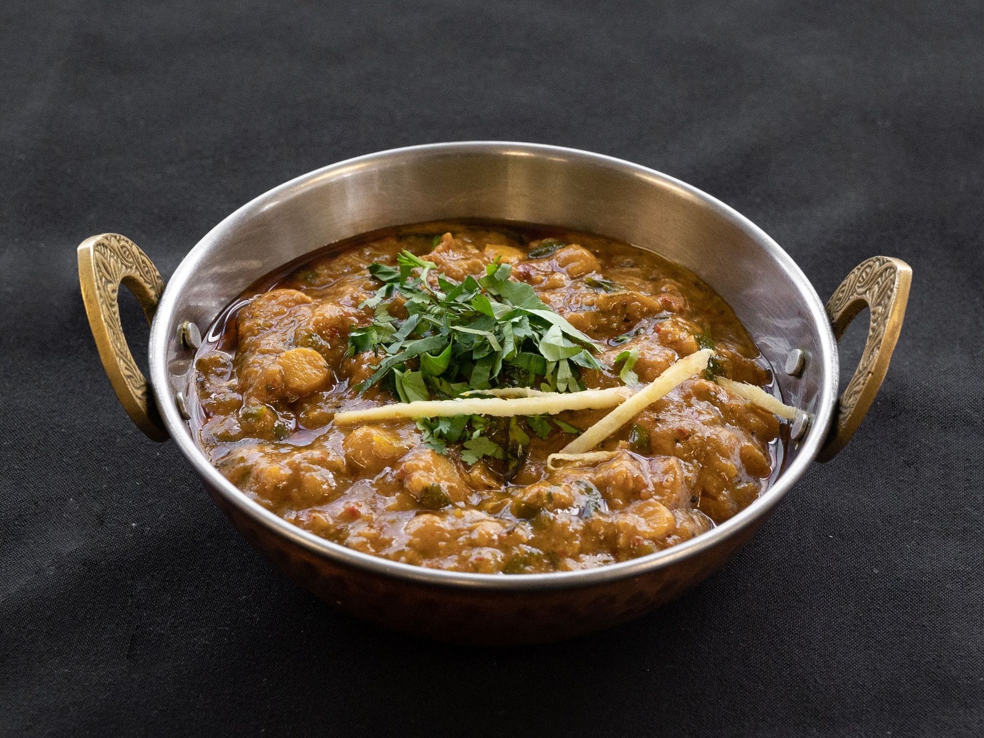 Chana masala in a copper bowl, garnished with cilantro and ginger, on a black surface.