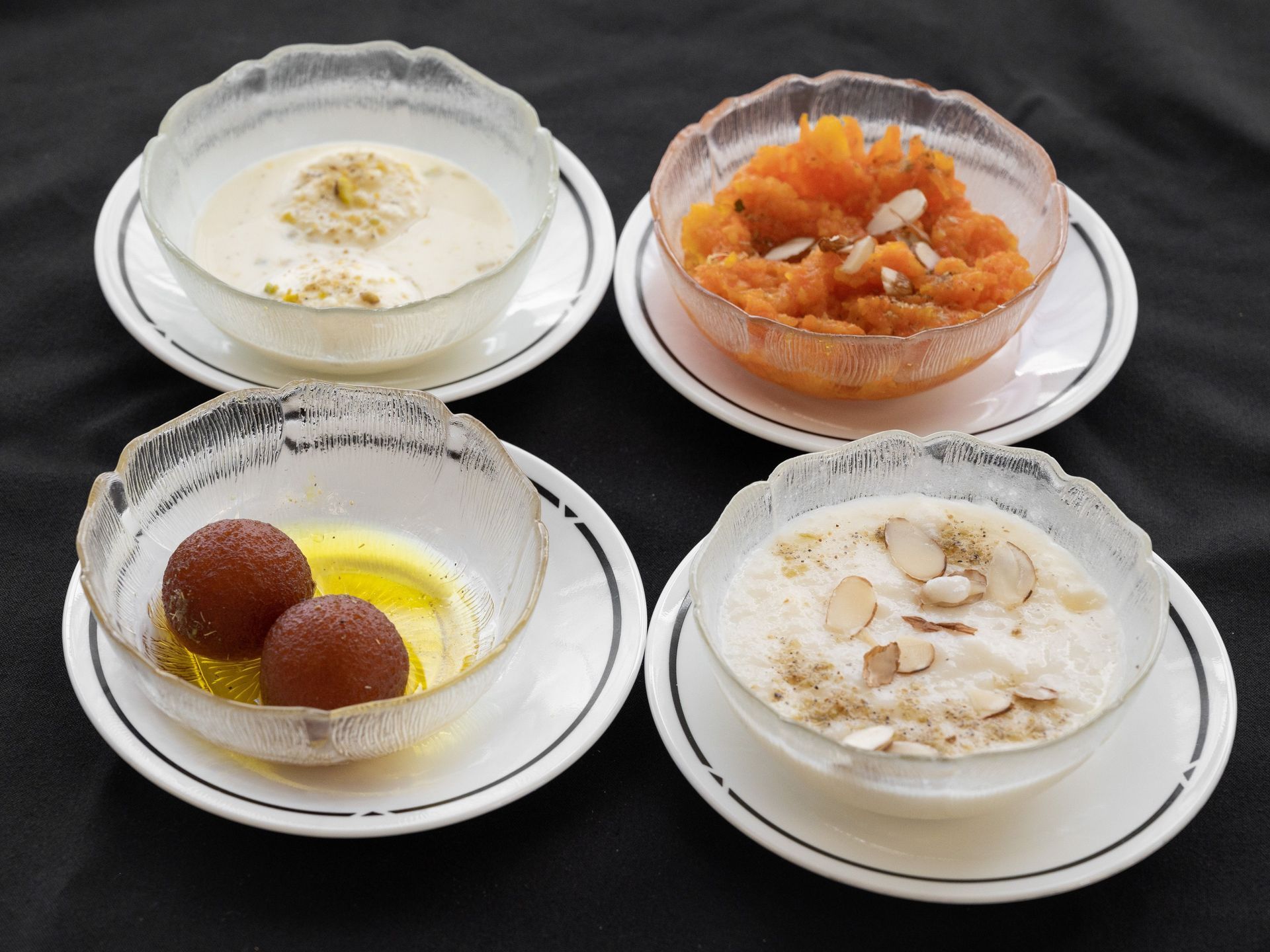 Four bowls of Indian desserts on saucers: rice pudding, carrot halwa, gulab jamun, and kheer.