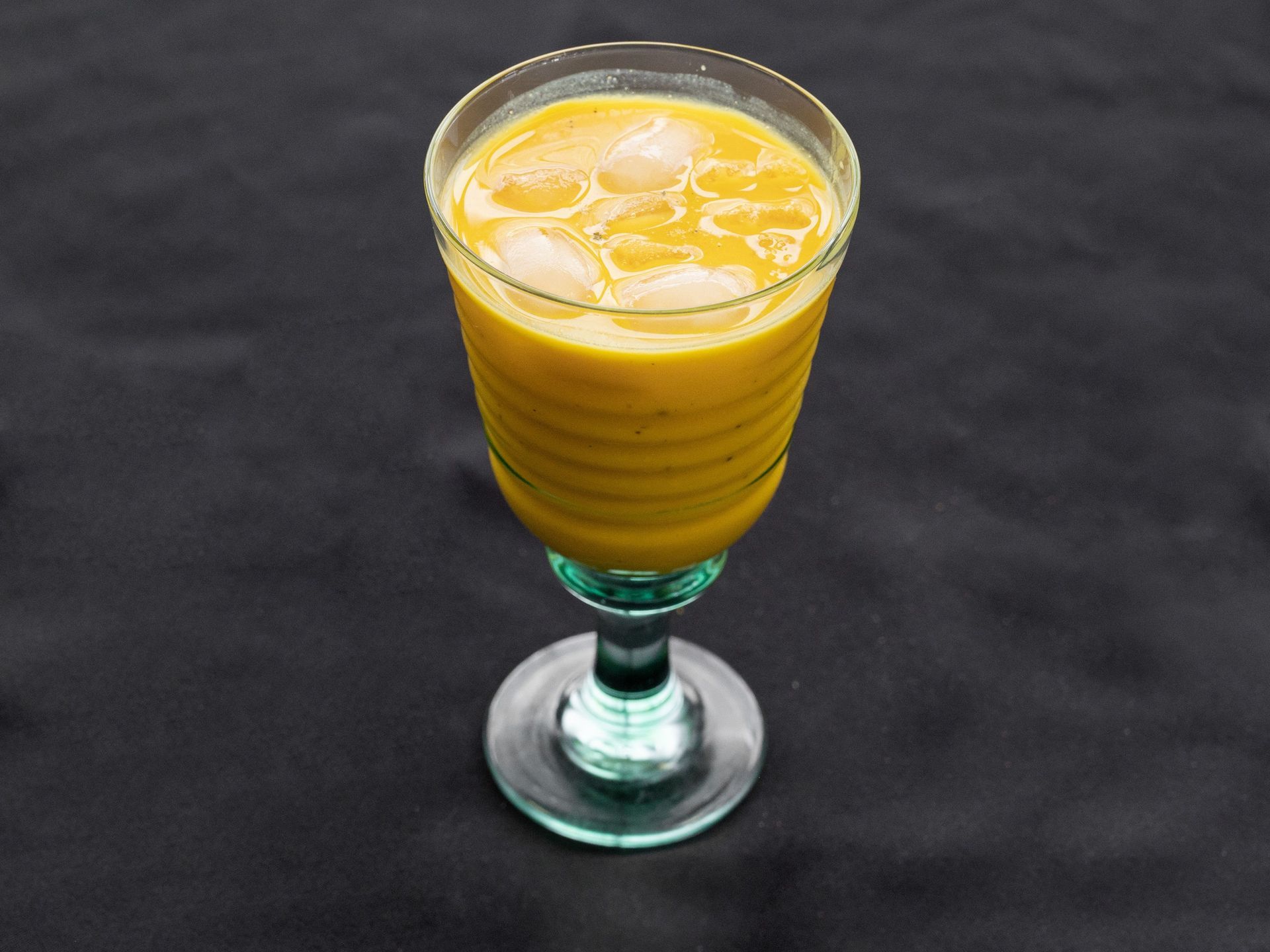 Yellow drink in a ridged glass with ice cubes on a dark surface.