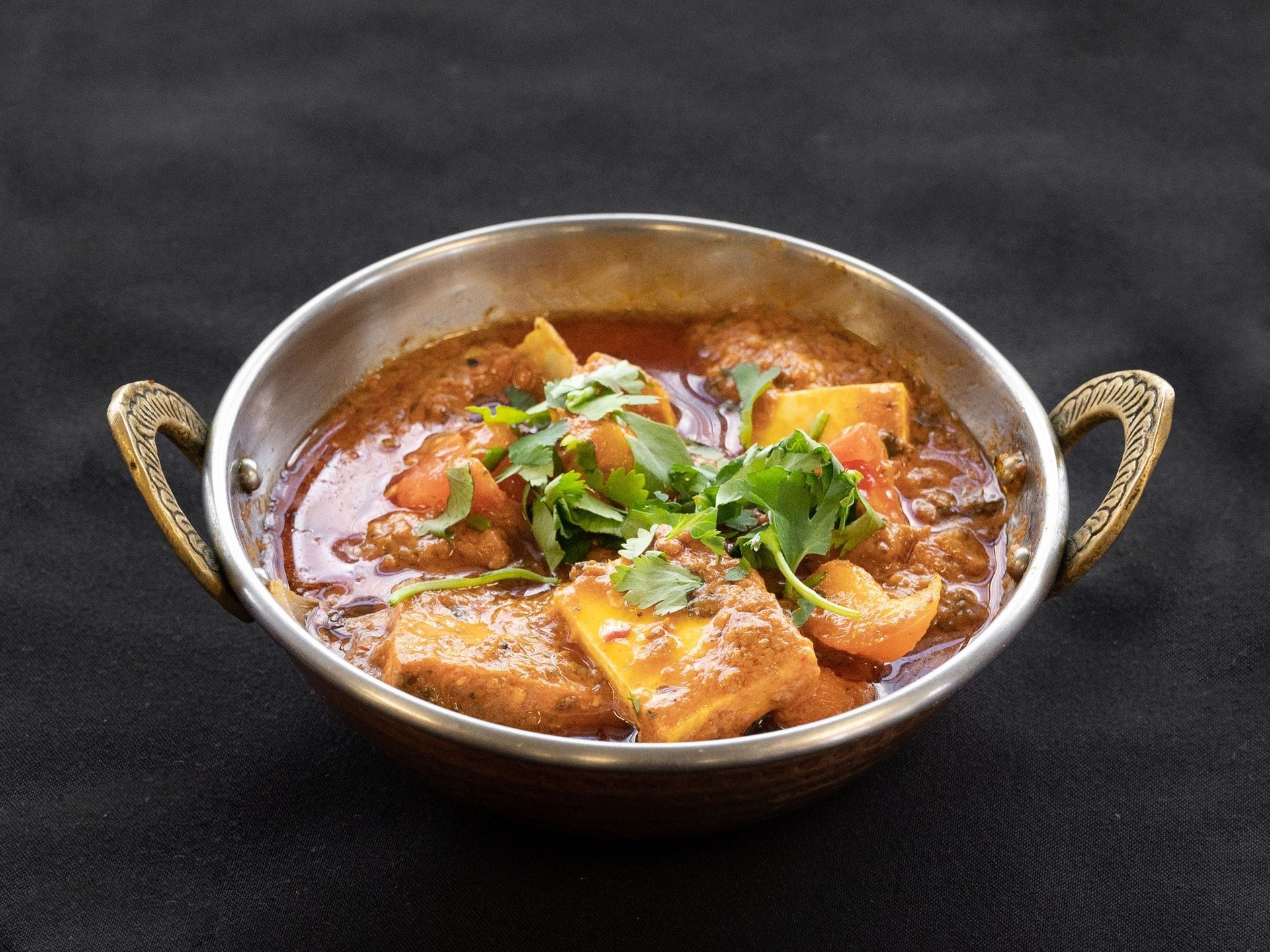 Paneer curry in a decorative metal bowl, garnished with cilantro, on a black surface.