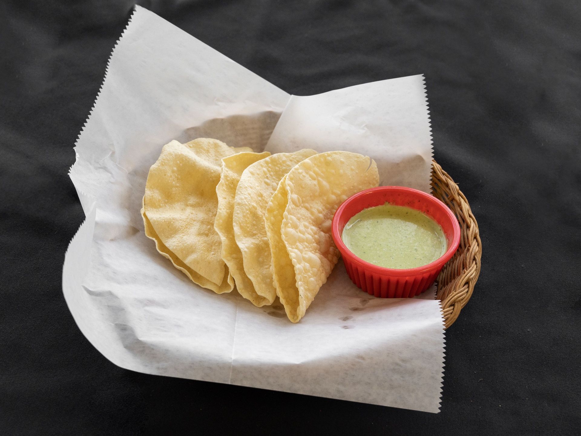 Crispy Indian papadums and green chutney in a basket, on a dark surface.