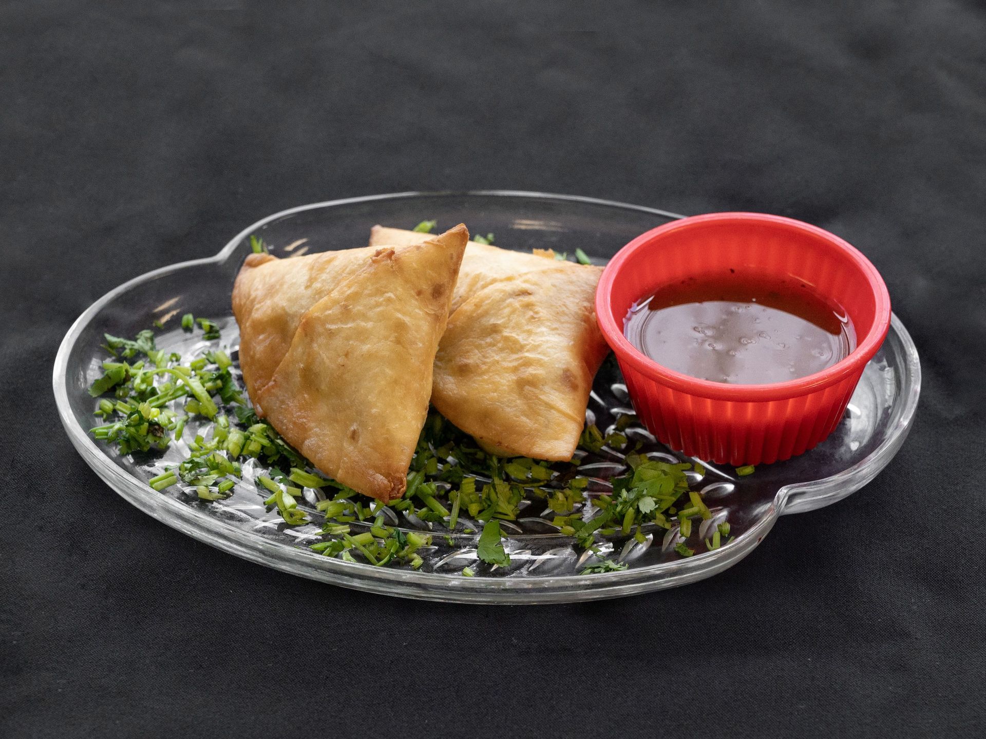 Two golden samosas with chutney on a clear plate garnished with chopped greens.